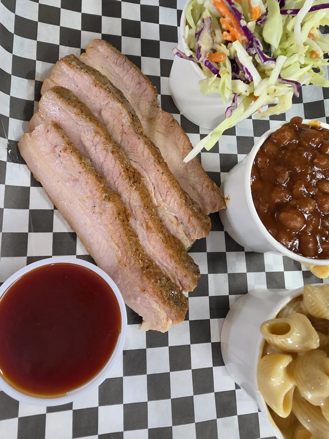 Sliced smoked brisket on checkered paper with BBQ sauce, baked beans, coleslaw and mac and cheese