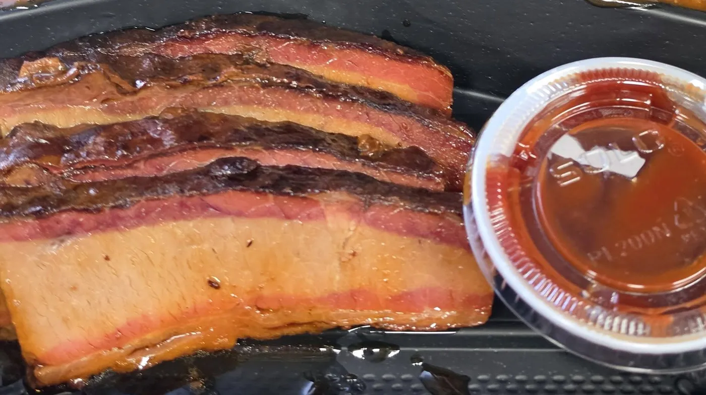 Slices of their smoked brisket showing a thick red-pink smoke ring under the bark, with a cup of house sauce